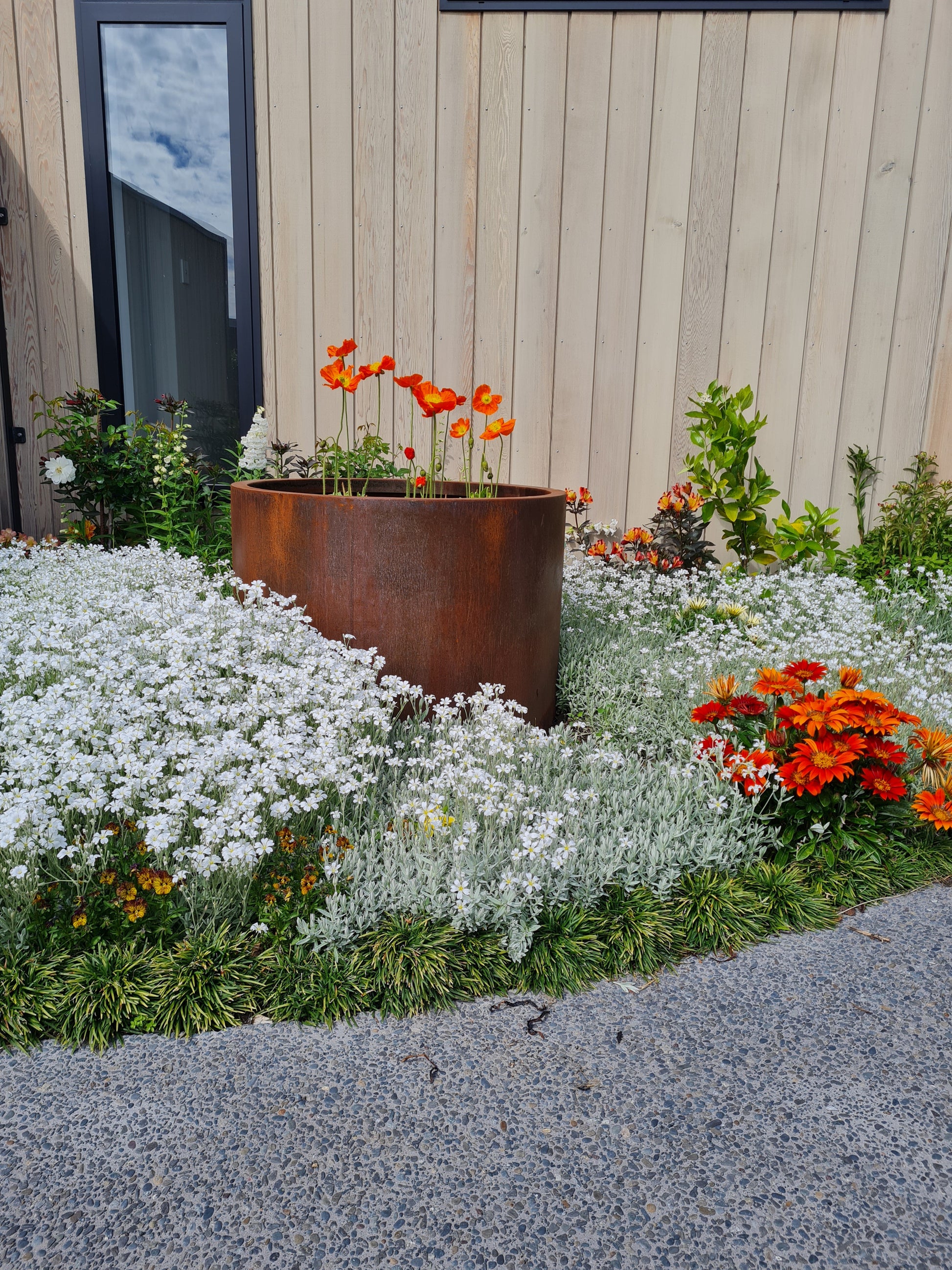 A rust-colored Corten cylindrical planter with plants, placed on a garden bed with various other plants around it.