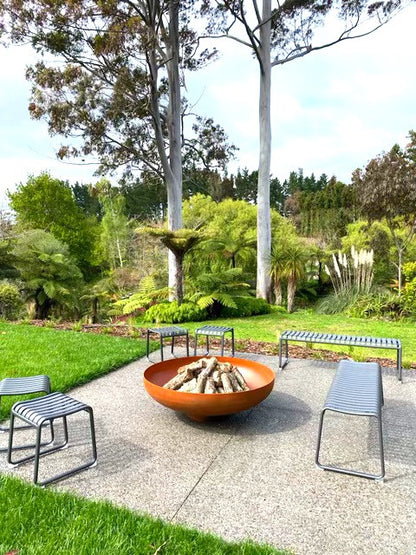 A large cylindrical corten steel fire bowl placed on a concrete surface with seating arrangements around it. The bowl appears to have a fire or charcoal-like substance at the bottom.