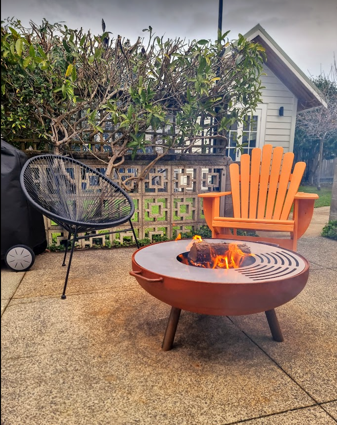 Outdoor fire bowl called The Whitianga with a cooking top, placed on a patio next to an orange chair and a white house in the background.